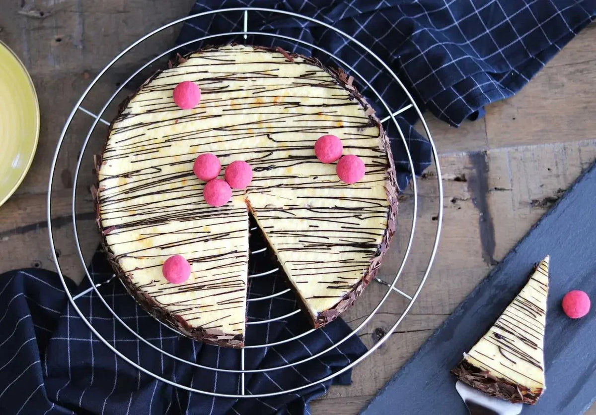 Käsekuchen mit Brownieboden, dekoriert mit Schokoladenstreifen und rosa Kugeln, auf einem Gitter mit Serviette.