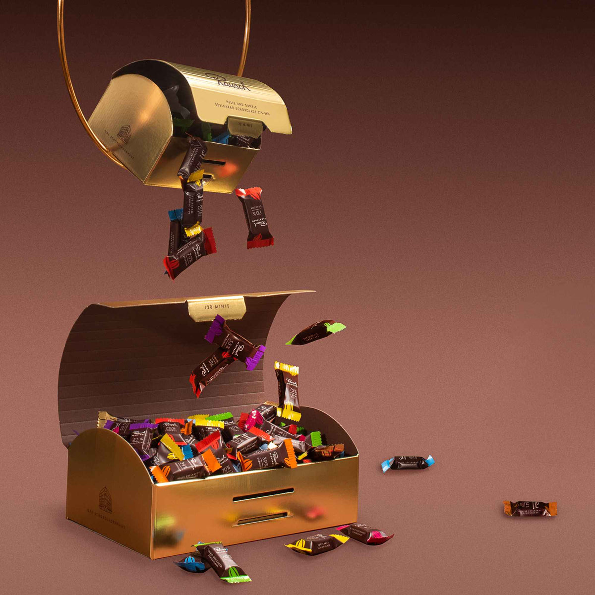Geöffnete goldene Box mit bunten Rausch Schokoladen-Minis, die herausfliegen; luxuriös und einladend präsentiert.