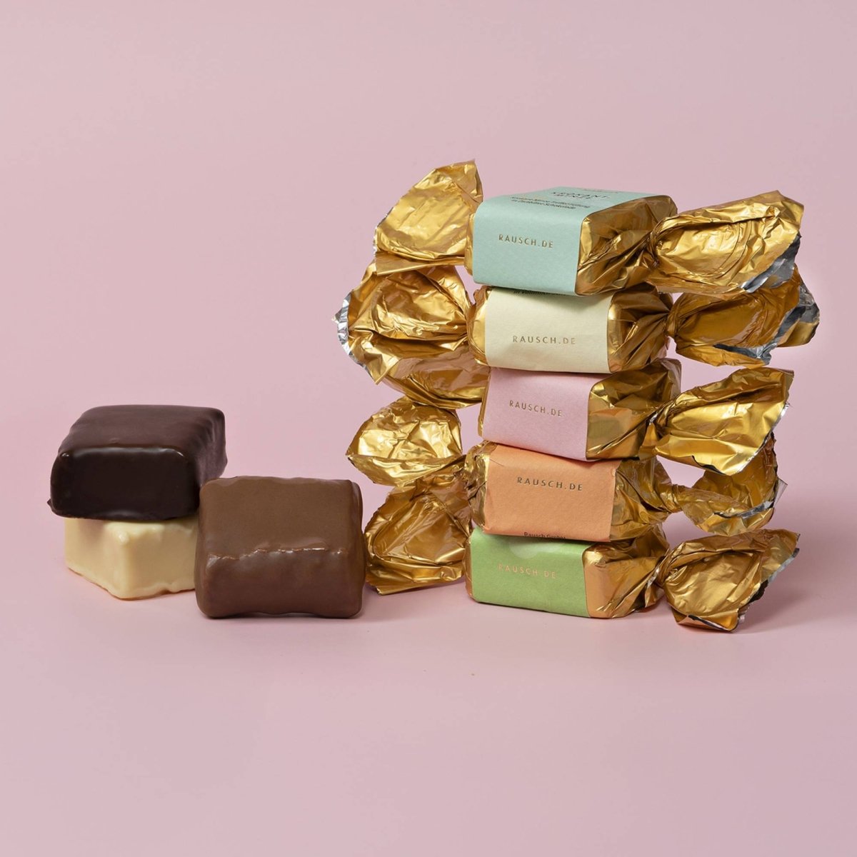 Mehrfarbige Happen in goldener Folie und zwei Schokowürfel mit Zuckerglasur auf rosa Hintergrund.