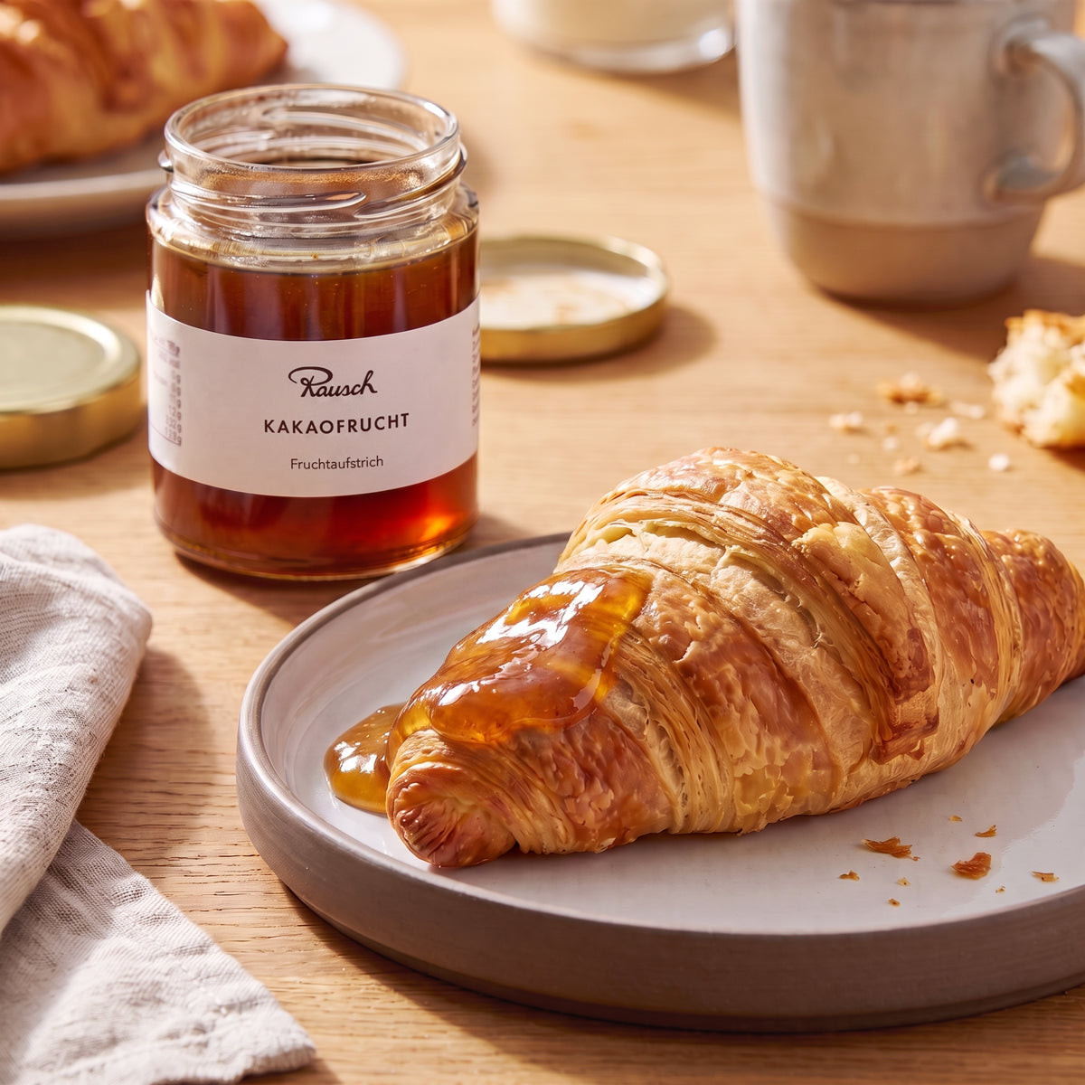 Glänzender Kakaofrucht-Aufstrich in Glas neben goldbraunem Croissant auf grauem Teller. - Rausch