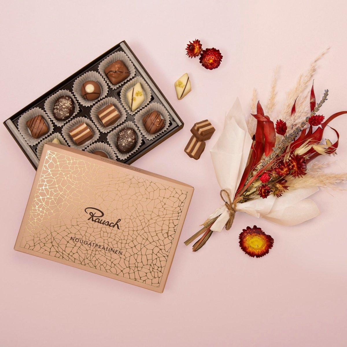 Rausch Pralinenbox mit neun Pralinen neben einem Trockenblumenstrauß auf rosa Hintergrund.