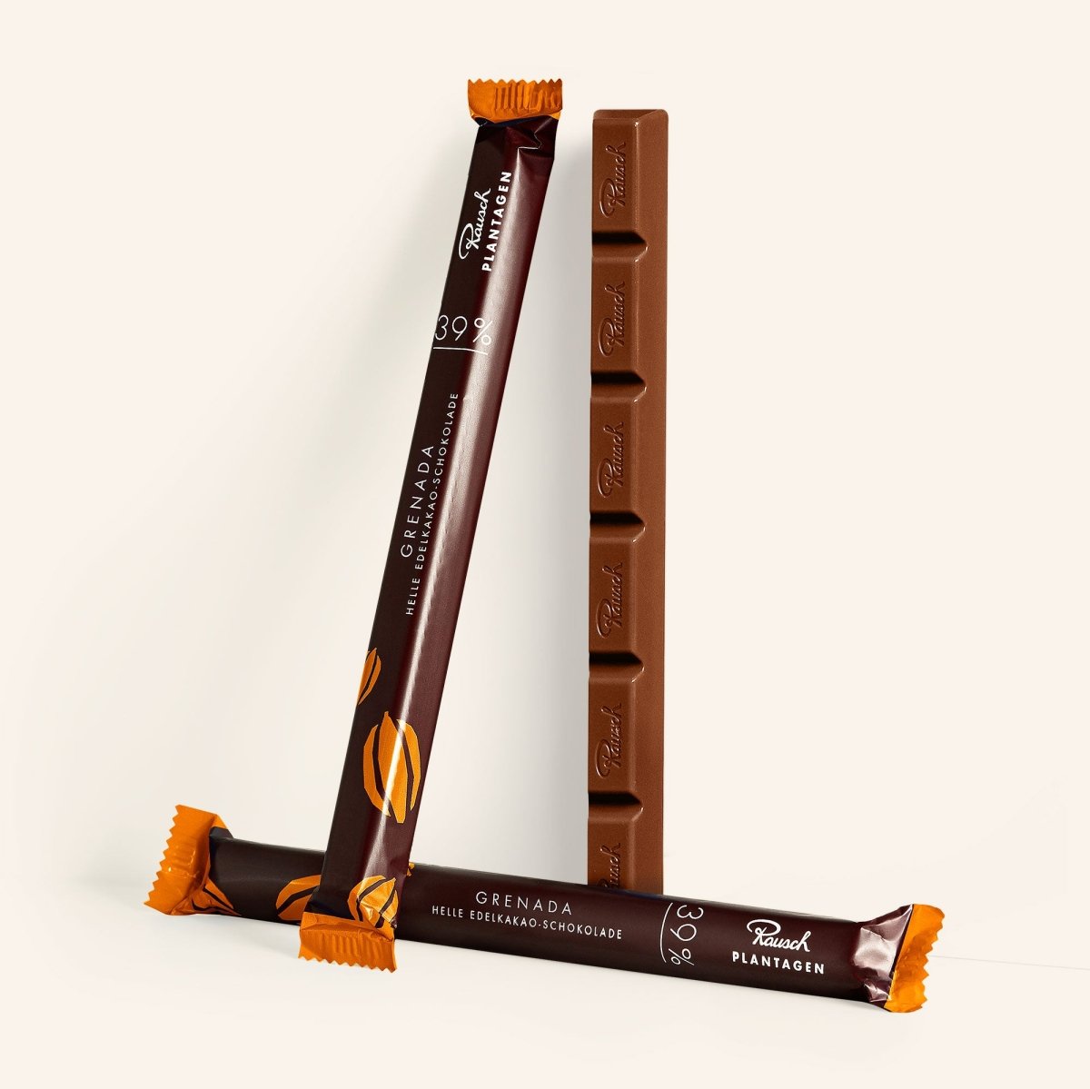 Drei Riegel Grenada Helle Edel-Kakaoschokolade, zwei stehend, einer liegend, mit brauner Verpackung und orangenen Akzenten.