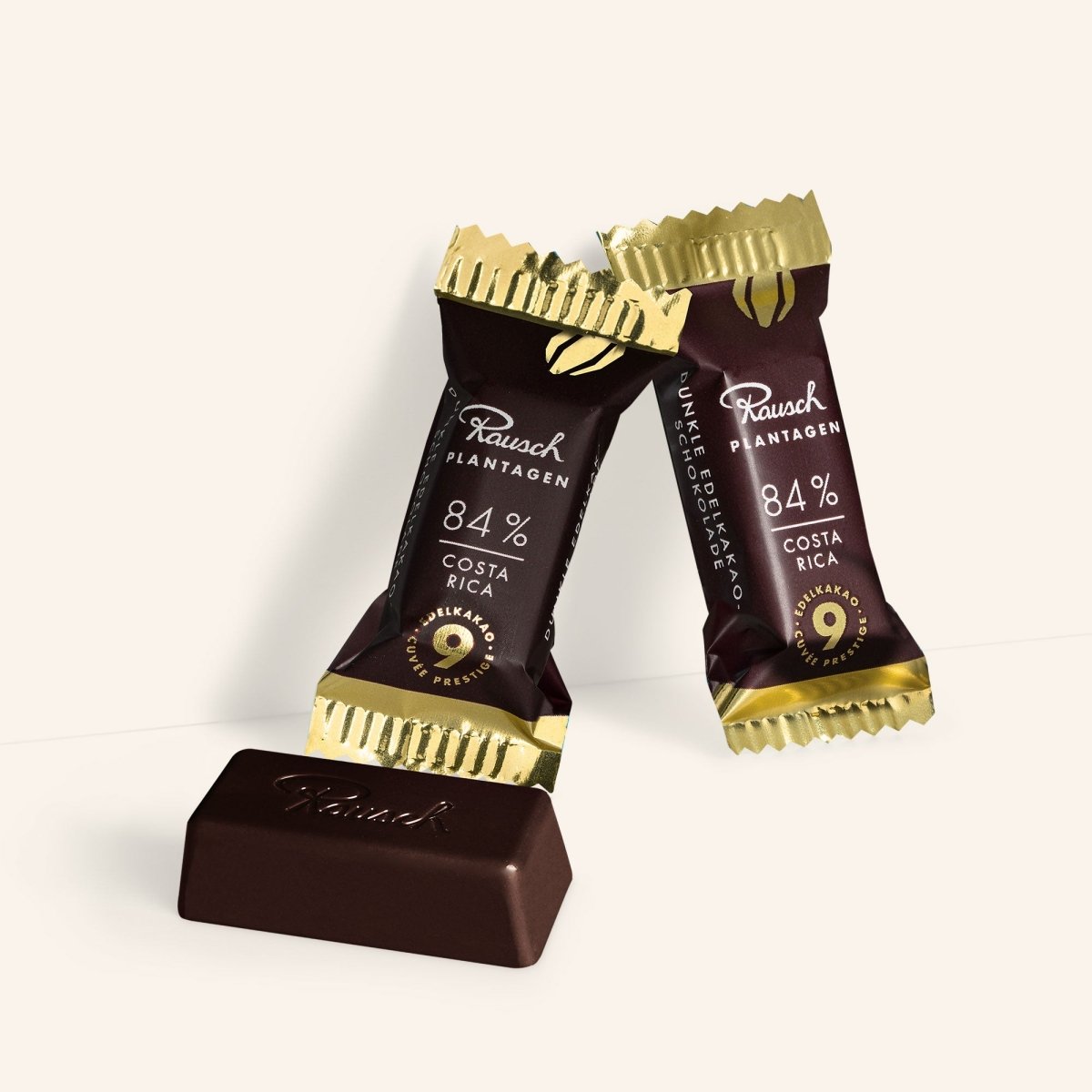 Zwei Rausch Costa Rica 84% Schokoladenminis vor hellem Hintergrund, einer ausgepackt, golden-braune Verpackung.