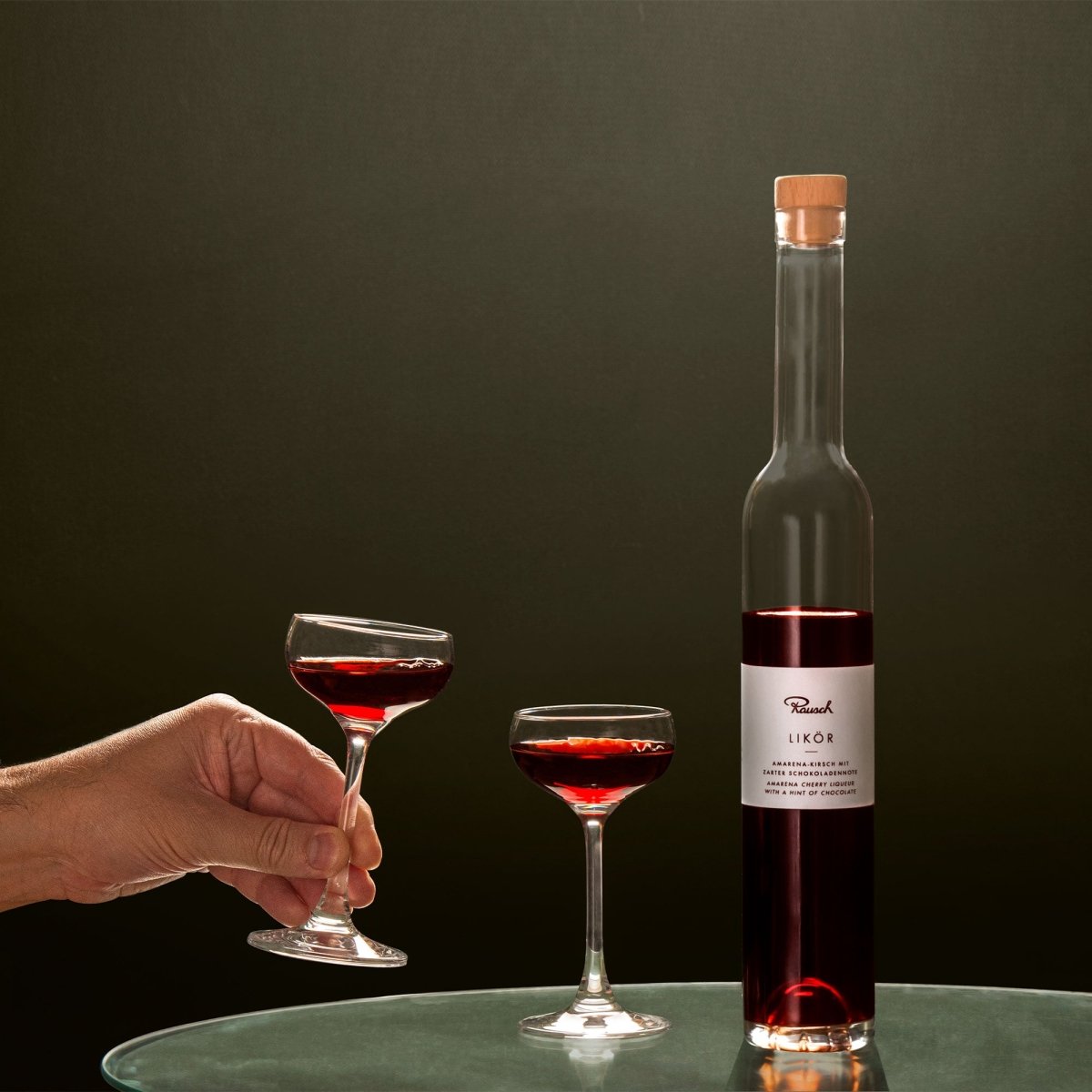 Zwei Gläser und eine Flasche roter Likör auf einem Tisch, eine Hand hält ein Glas an.