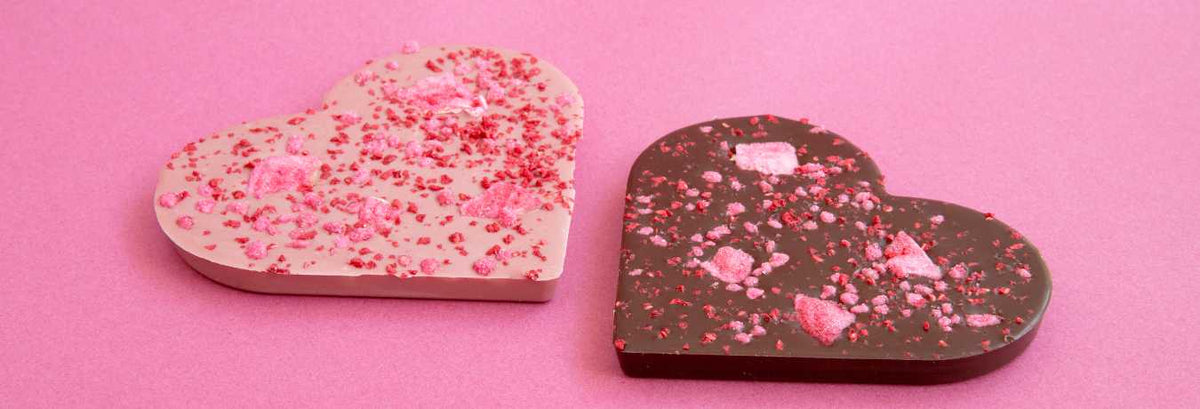 Zwei herzförmige Schokoladenstücke, eins weiß mit rosa Streuseln, das andere dunkel mit roten und rosa Streuseln, auf rosa Hintergrund.