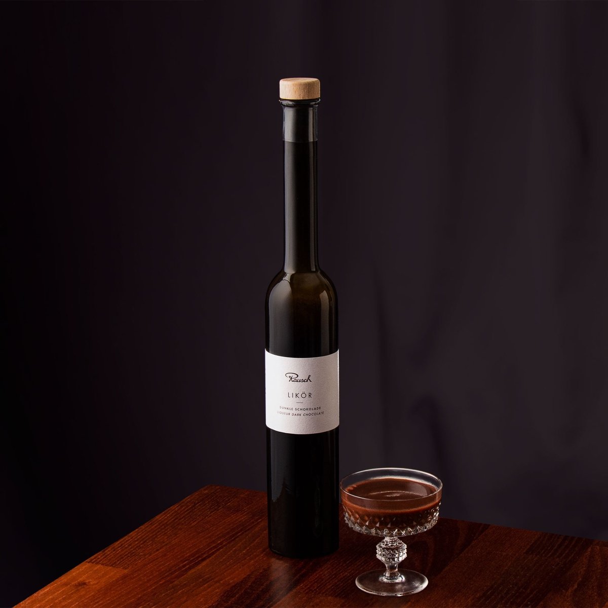 Elegante Flasche dunklen Likörs neben einem Glas auf einem Holztisch vor schwarzem Hintergrund.