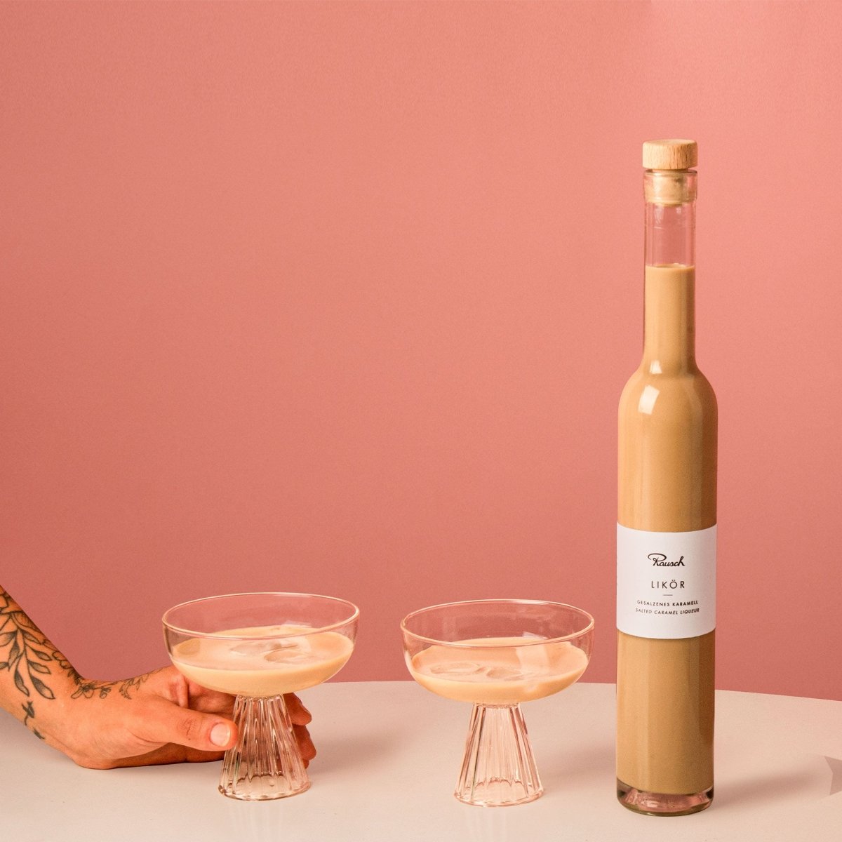 Flasche gesalzener Karamell-Likör mit zwei Gläsern, vor rosa Hintergrund, Hand greift nach einem Glas auf weißem Tisch.
