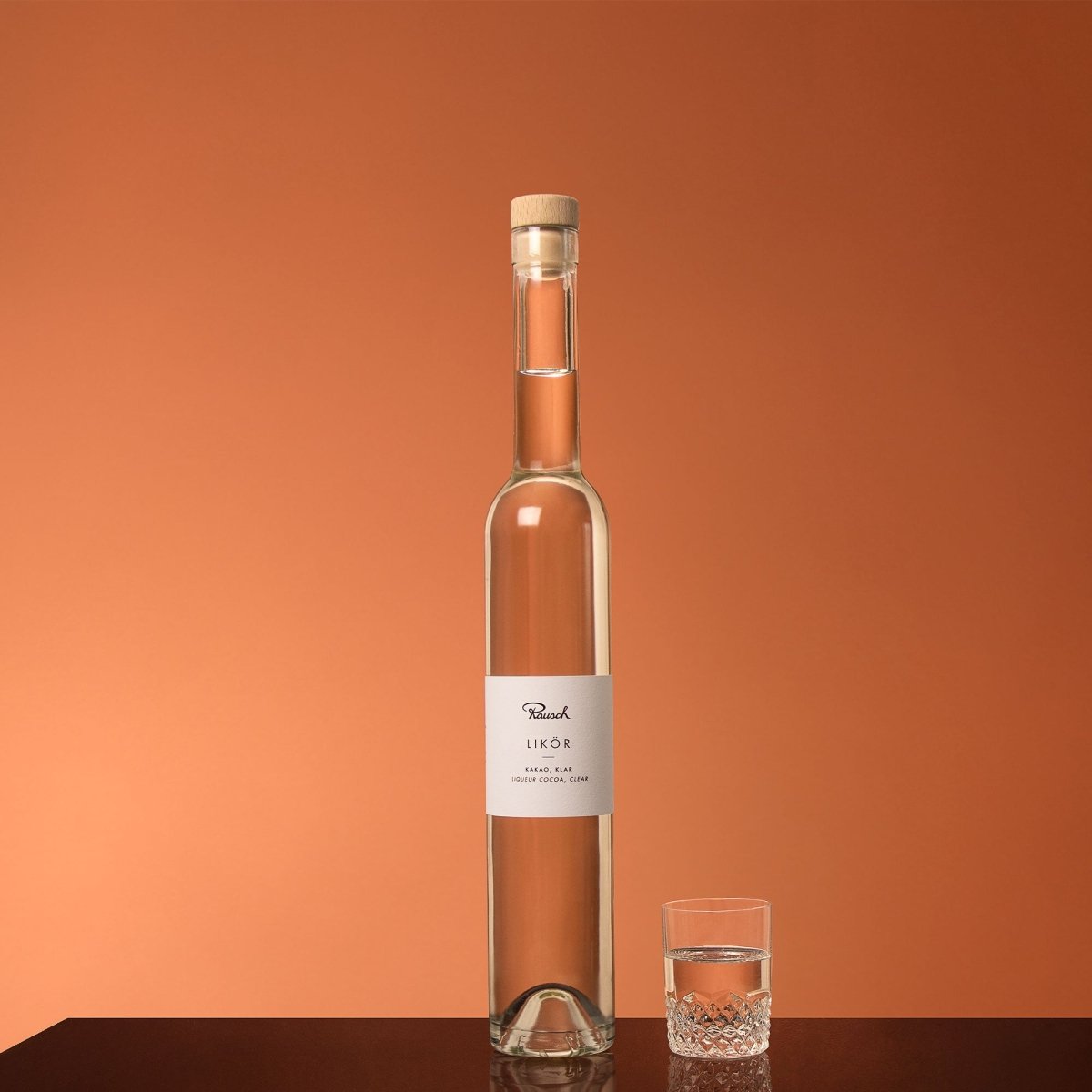 Klarer Kakao-Likör in schmaler Flasche mit Glas auf Holzoberfläche vor warmem orangem Hintergrund.