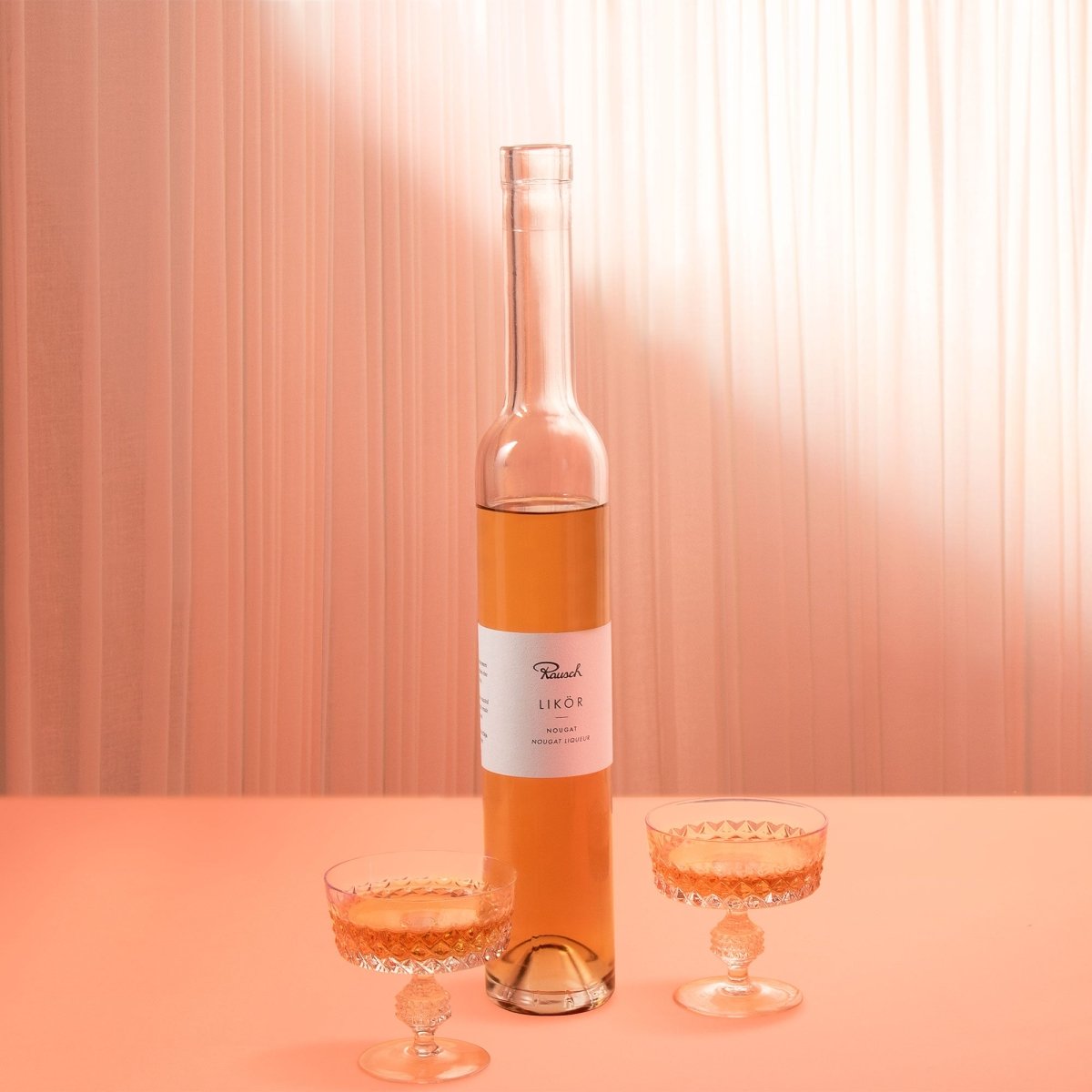Flasche Rausch Nougatlikör mit zwei Gläsern auf rosa Hintergrund.