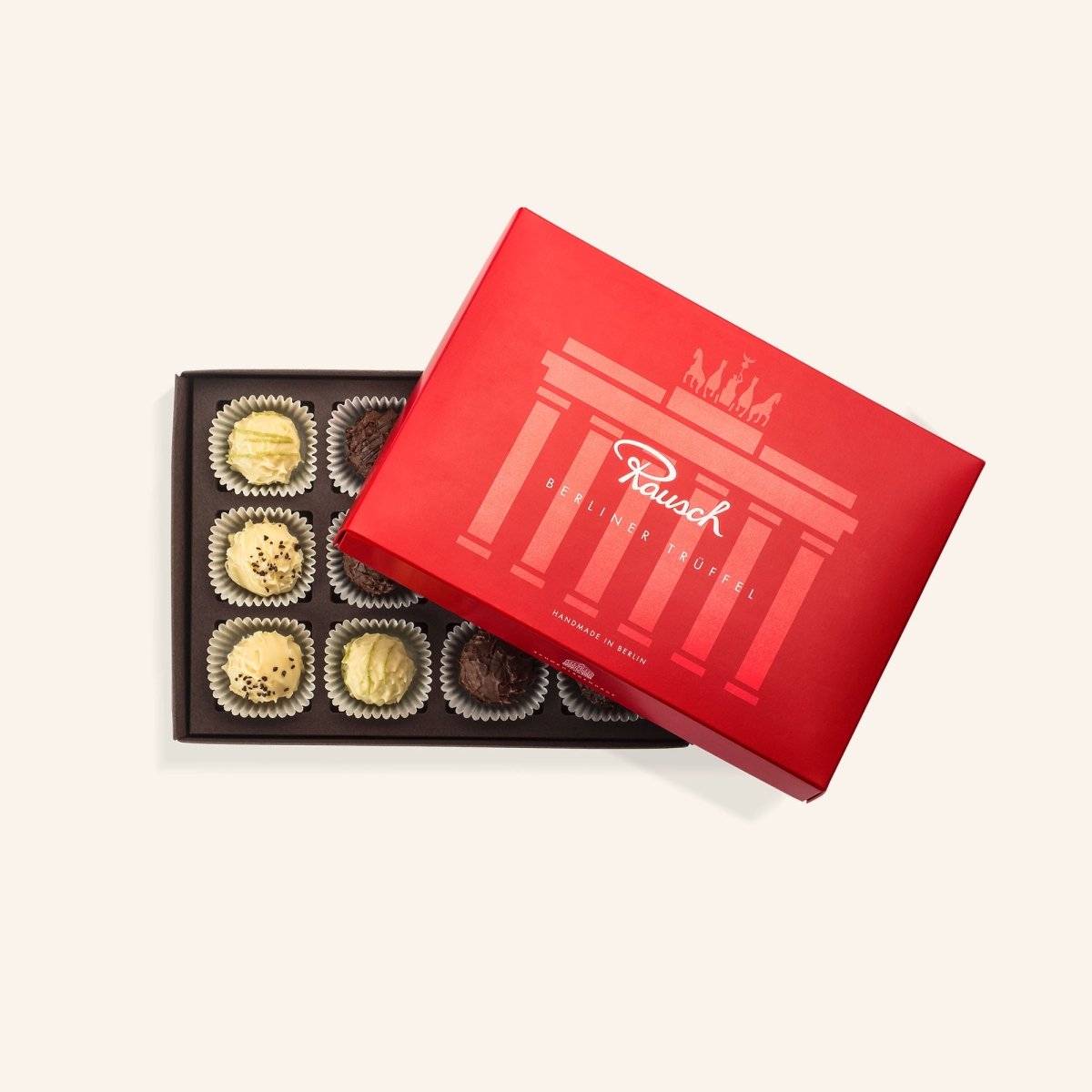 Rote Pralinenschachtel mit Berliner Trüffeln, teils in Goldfolie, geöffnet auf beigem Hintergrund.