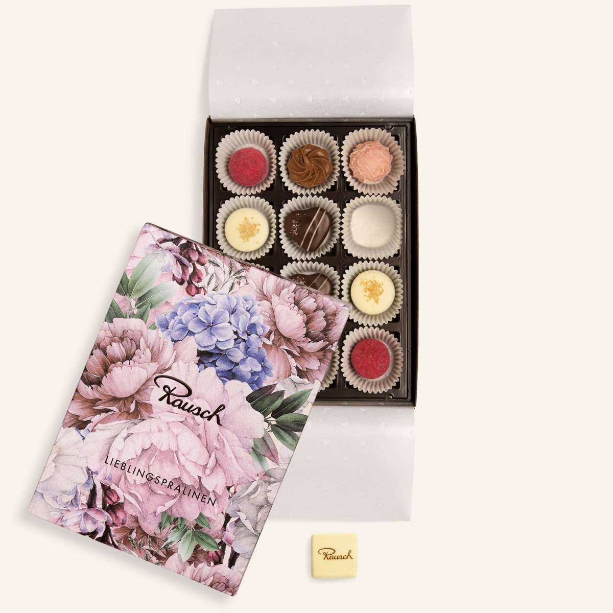 Offene Pralinenbox mit 12 sortierten Pralinen und Blumenmuster-Deckel, daneben weiße Rausch-Praline.