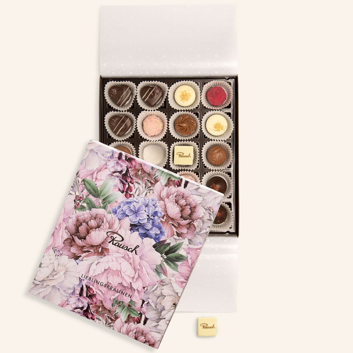 Geöffnete Schachtel mit 20 Pralinen, verziert mit Blumenmotiv, einzelne Praline daneben liegend.