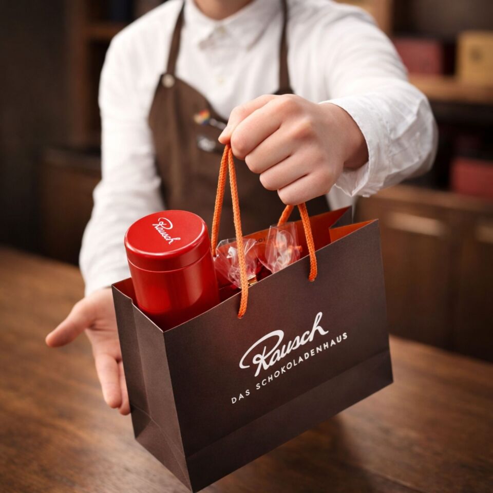 Hand überreicht Rausch-Schokoladenhaus-Tüte mit roter Dose und Pralinen auf Holztisch.
