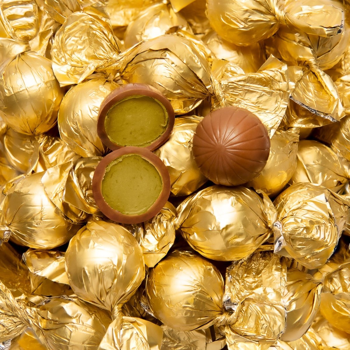 Goldene Pralinenkugeln und eine geöffnete Pralinenkugel mit grünem Kern, umgeben von edler Schokolade.