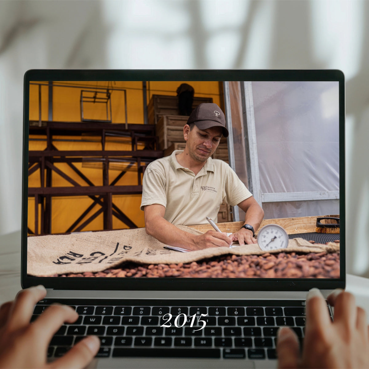 Person in Arbeitskleidung, überprüft Kakaobohnen auf einem Laptop-Bildschirm, dahinter Industriekulisse.
