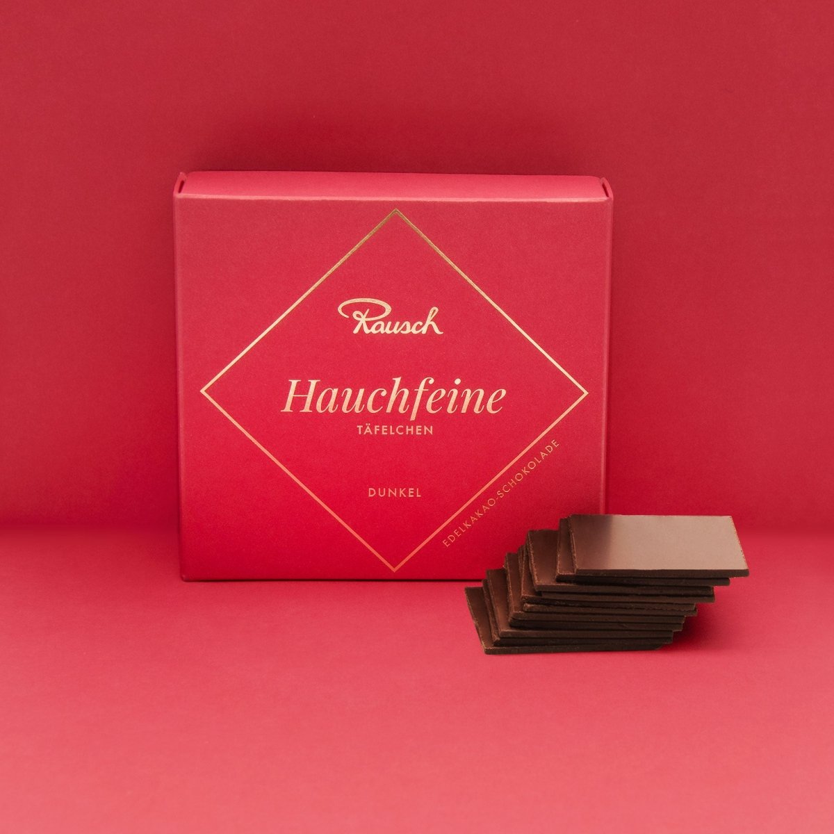 Rote Verpackung mit "Rausch Hauchfeine Dunkel" Schokolade neben dünnen Schokoladenstücken auf rotem Hintergrund.