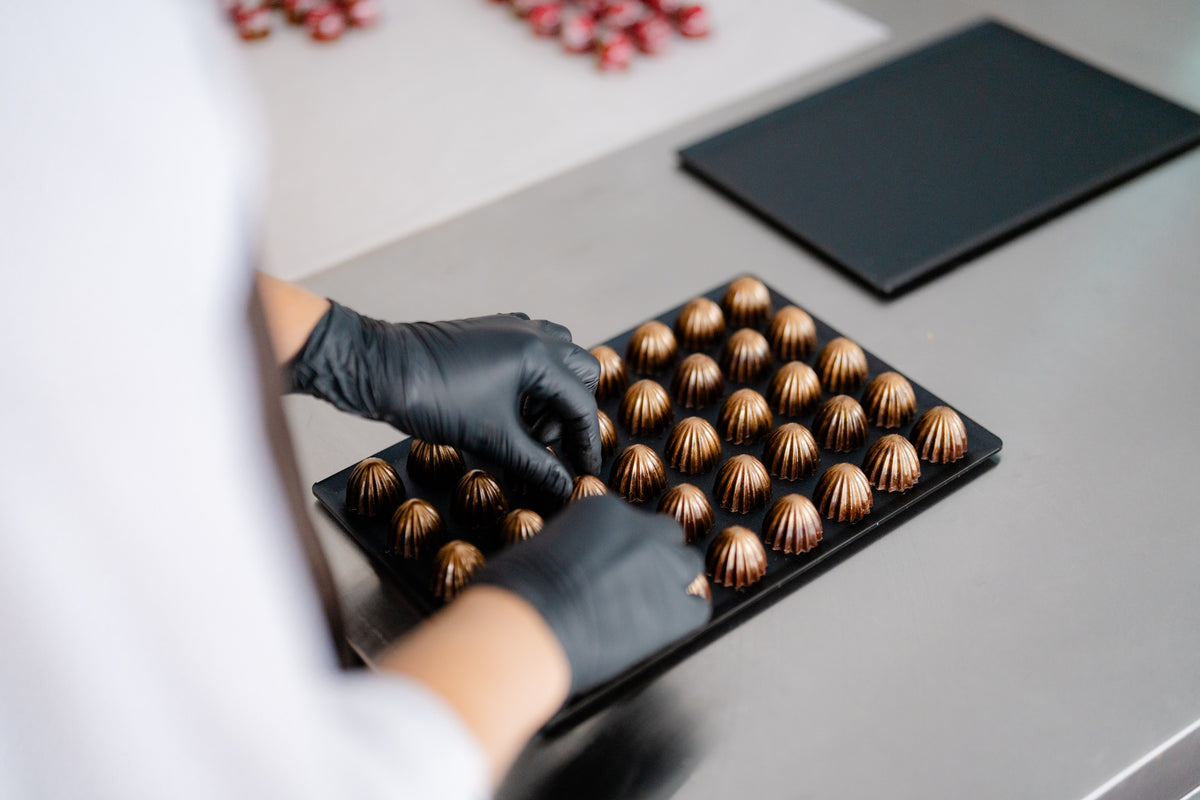 Hand in schwarzen Handschuhen arrangiert Pralinen auf einem Tablett in einer Patisserie.