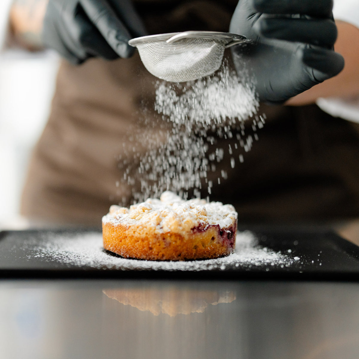 Person streut Puderzucker auf kleinen Kuchen auf schwarzem Teller.