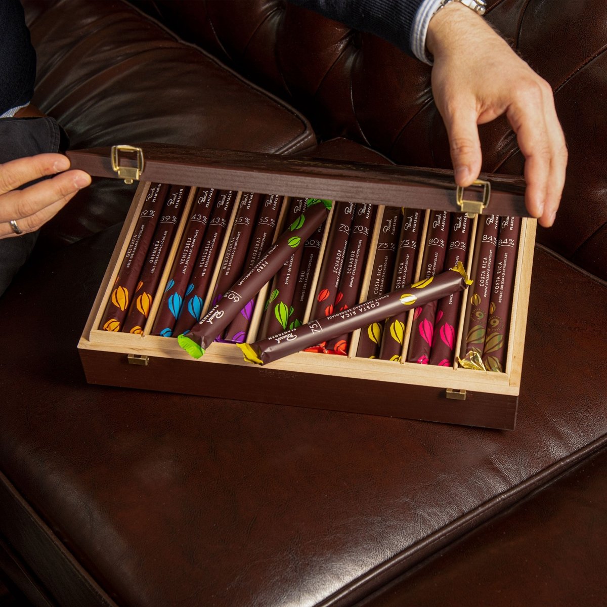 Holzbox mit Rausch Schokoladen-Sticks in verschiedenen Farben wird geöffnet, liegend auf braunem Ledersofa.