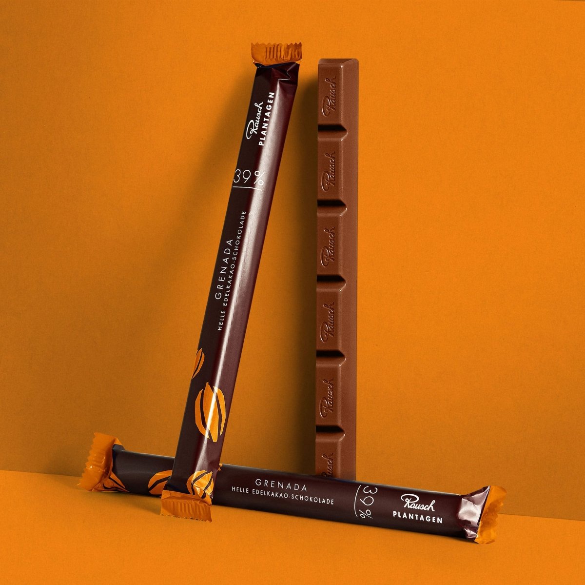 Zwei Schokoladenriegel in brauner Verpackung mit "Grenada 39" und heller Schokolade auf orangefarbenem Hintergrund.