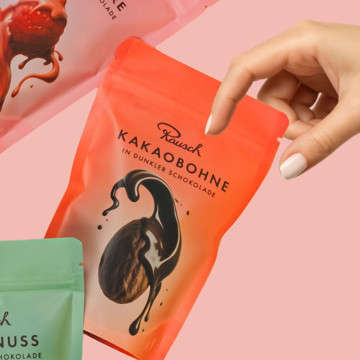 Hand greift nach roter Tüte mit dunklen Schokoladen-Kakaobohnen von Rausch vor rosa Hintergrund.