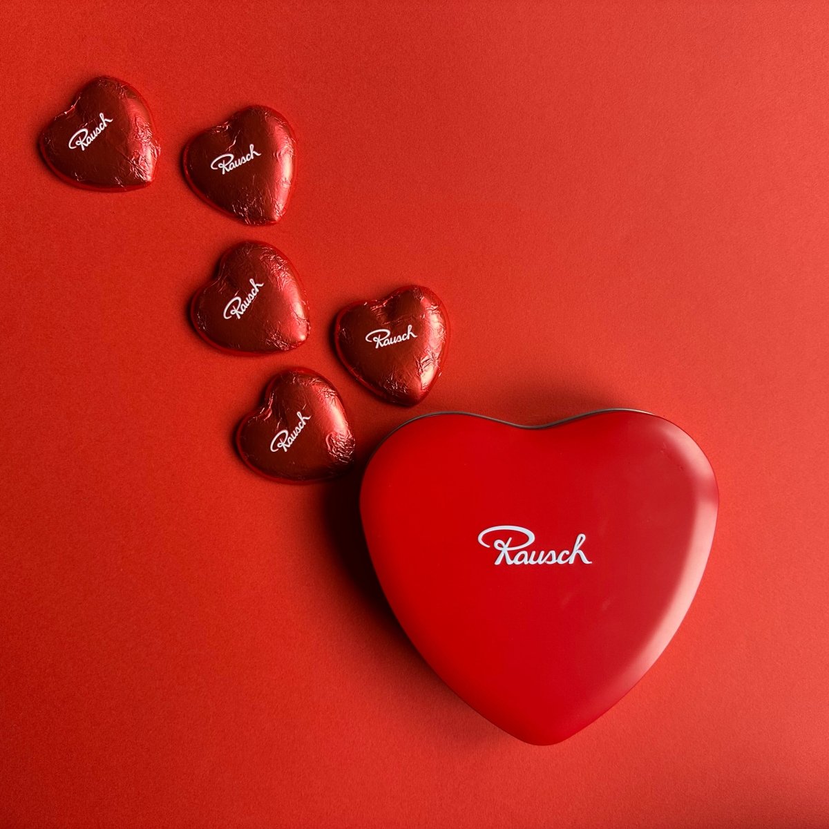 Rote herzförmige Pralinenschachtel und Schokopralinen von Rausch auf rotem Hintergrund.