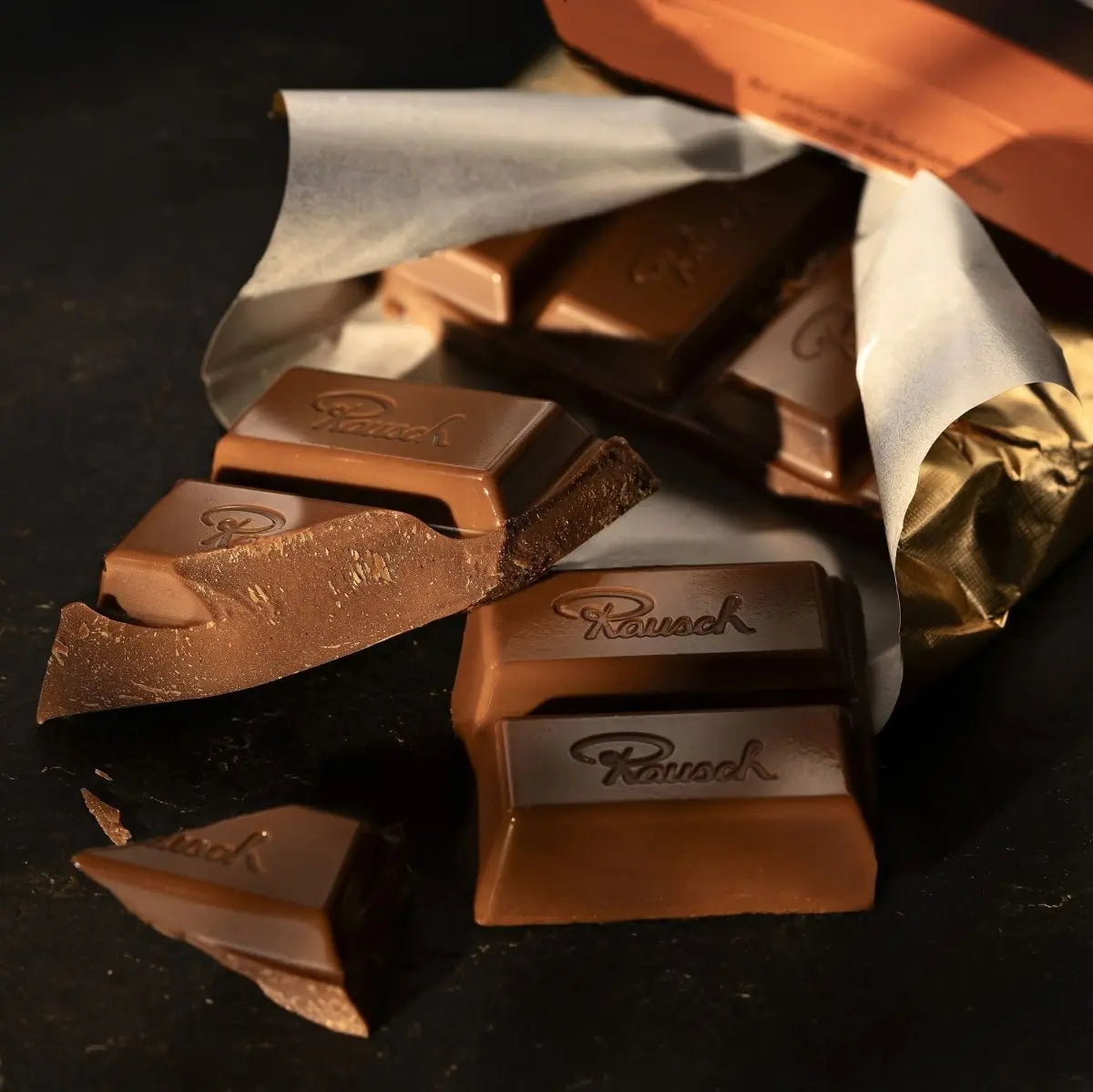Edelvollmilchschokolade von Rausch aus Verpackung mit goldener Folie, stückweise gebrochen auf dunklem Untergrund.