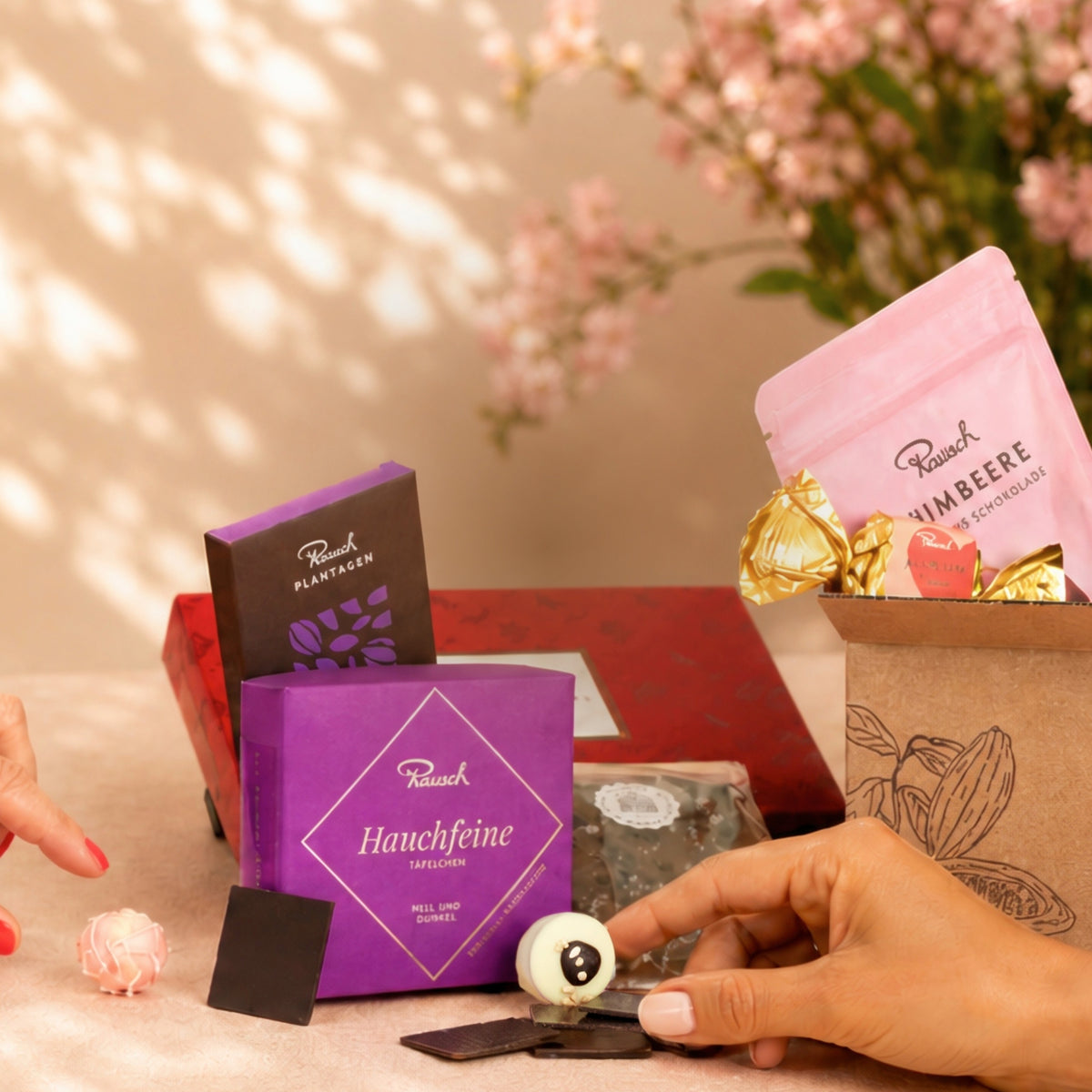 Personen arrangieren Rausch-Schokolade und Pralinen mit floraler Dekoration im Hintergrund.