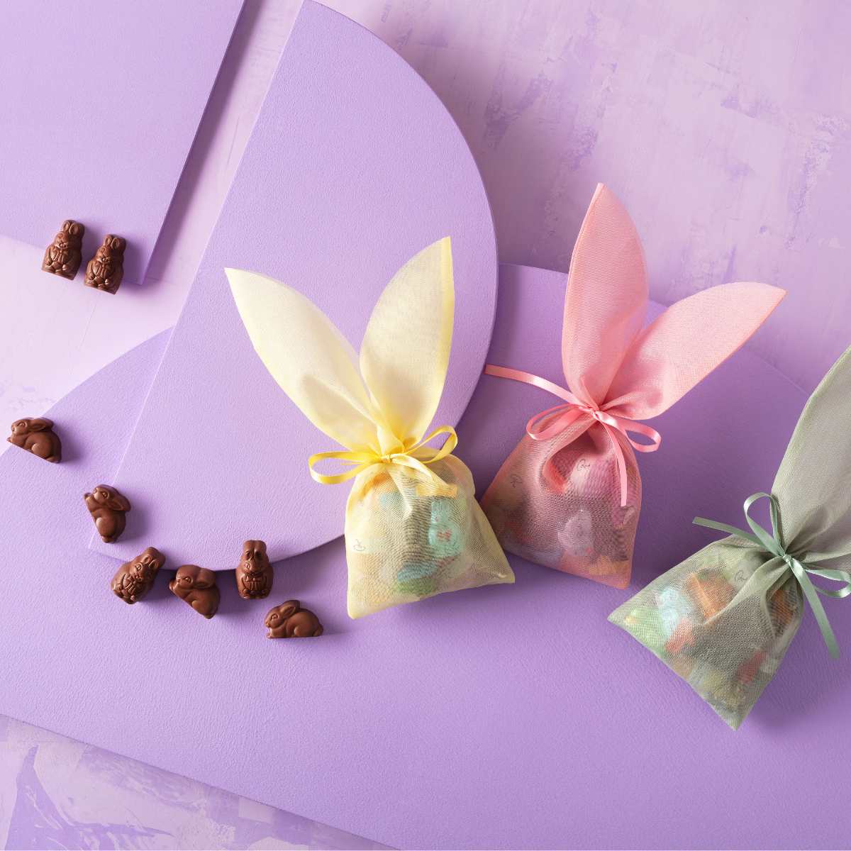 Osterbeutel mit Schokolade, umgeben von Schokohasen, auf lila Hintergrund arrangiert.