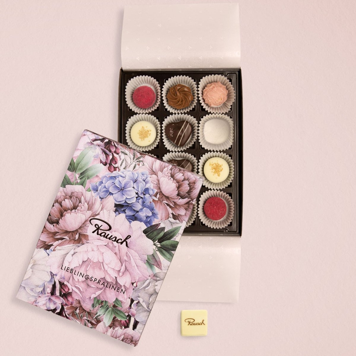 Box mit zwölf handgefertigten Pralinen, Rausch-Logo auf floralem Deckel, eine Praline daneben auf rosa Hintergrund.