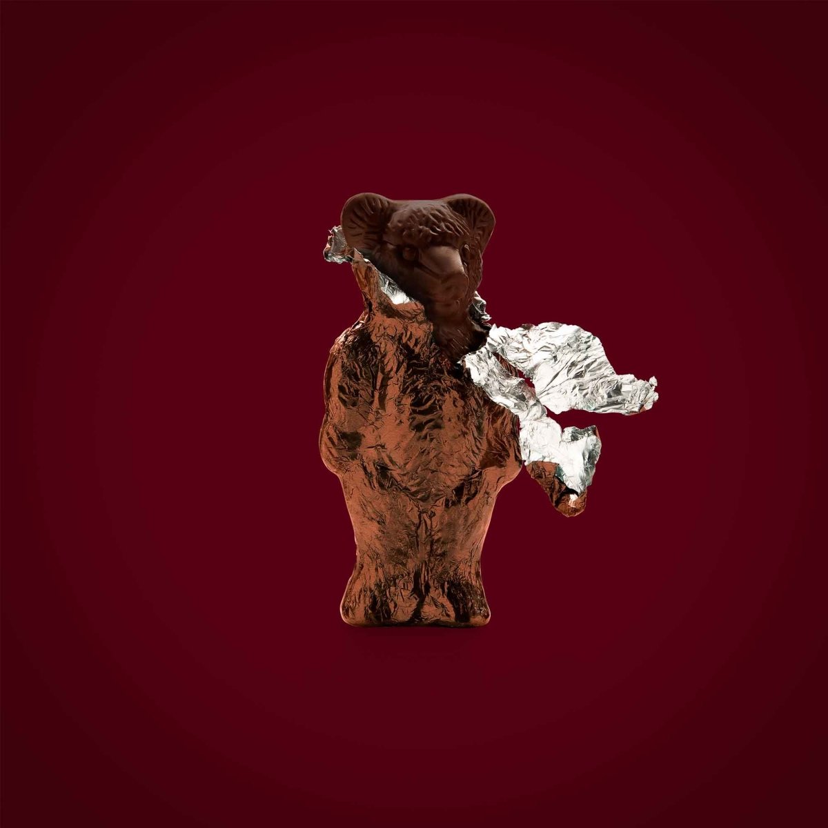 Schokoladenbär aus brauner Rausch-Schokolade mit Folie, vor dunkelrotem Hintergrund.