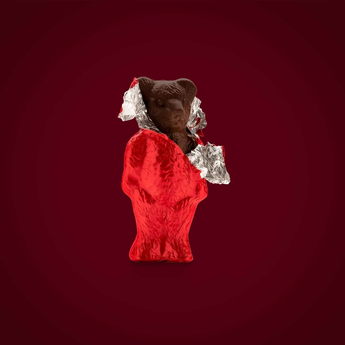 Schokoladenbär in roter Folie, teils ausgepackt, vor dunkelrotem Hintergrund.