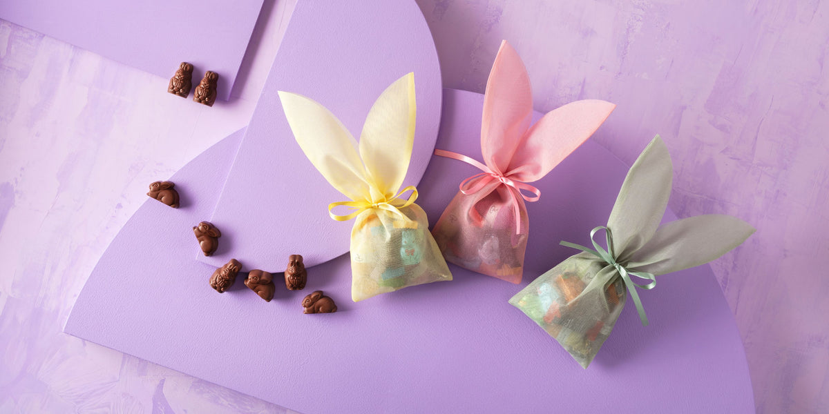 Drei bunte Hasenohrenbeutel mit Pralinen auf lila Hintergrund, umgeben von kleinen Schokoladenhasen.