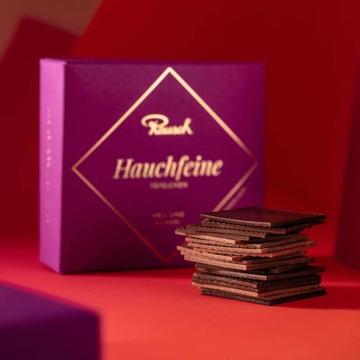 Stapel dünner Schokoladentafeln vor einer violetten Verpackung mit goldener Aufschrift "Hauchfeine Täfelchen" auf rotem Hintergrund.