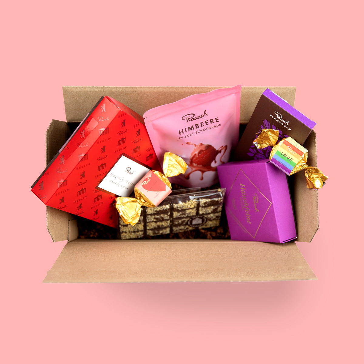 Offene Box mit Rausch-Schokoladen, Pralinen und einer Himbeer-Tafel auf rosa Hintergrund.